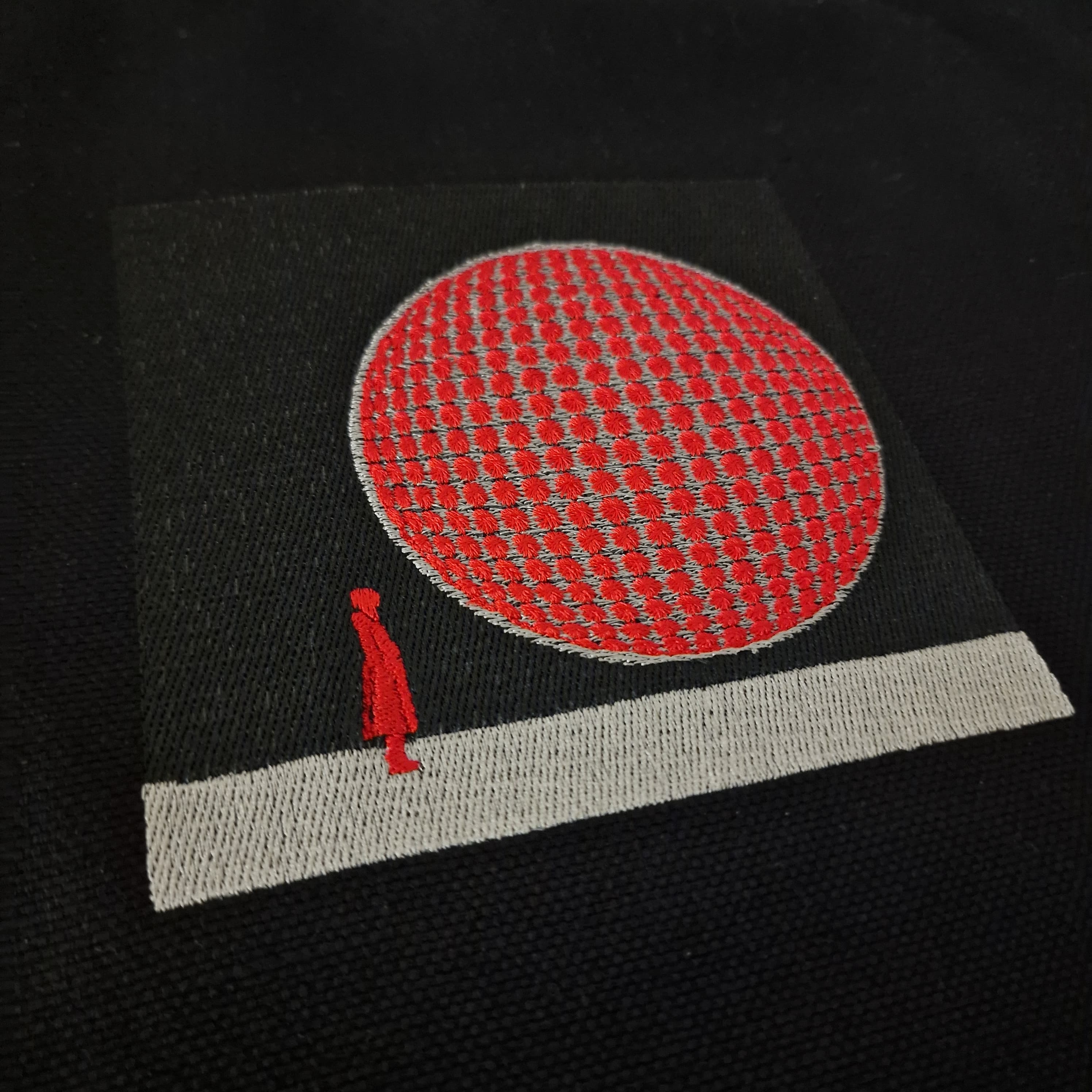 Embroidered design on a black fabric with a red circle and small figure, against a dark background.