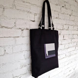 Black tote bag with geometric grey embroidery, hanging on a white brick wall