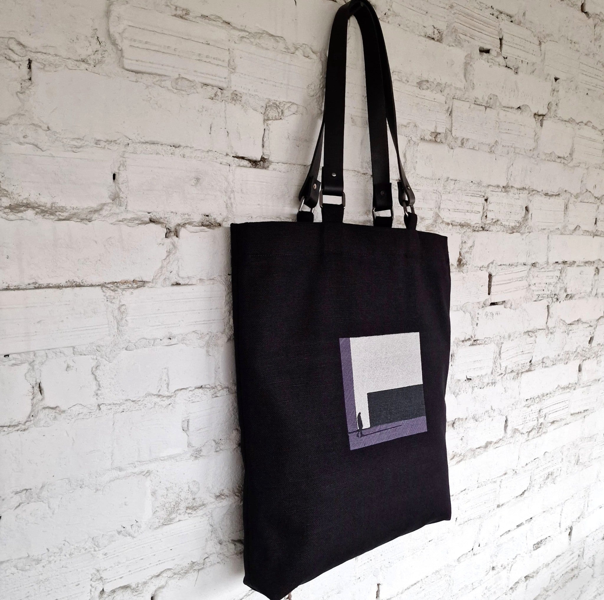 Black tote bag with geometric grey embroidery, hanging on a white brick wall