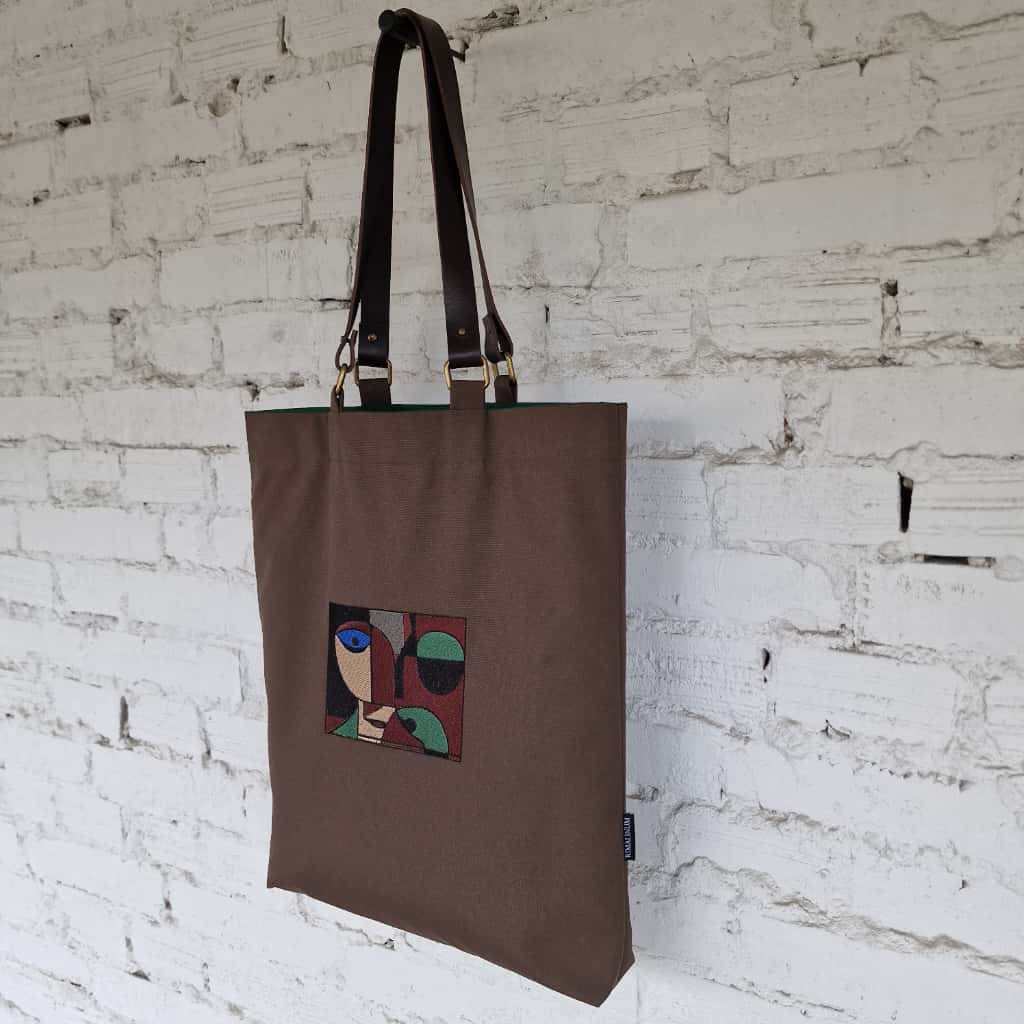 Dark brown cotton tote bag with embroidered abstract face design, leather handles, and green lining – handmade by RIMALINUM in Lithuania