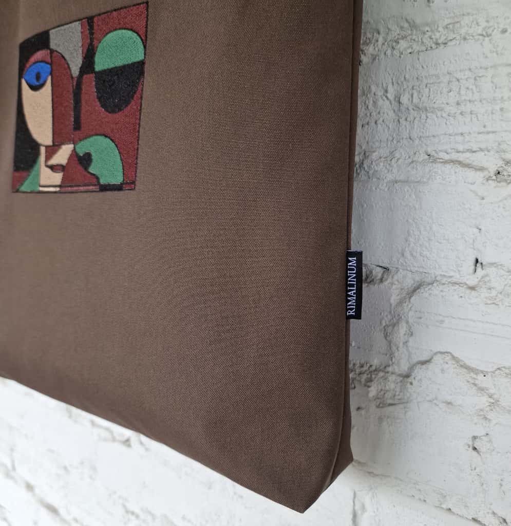 Close-up of abstract embroidered face design and RIMALINUM fabric label on dark brown cotton tote bag