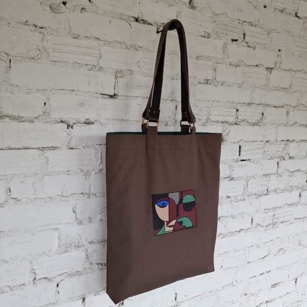 Dark brown cotton tote bag with embroidered abstract face design, leather handles, and green lining – handmade by RIMALINUM in Lithuania
