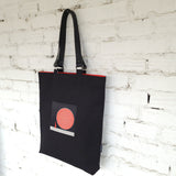 Black tote bag with a red and white design on a white brick wall background