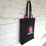 Black linen tote bag with 3D globe embroidery and leather handles, hanging against a white brick wall