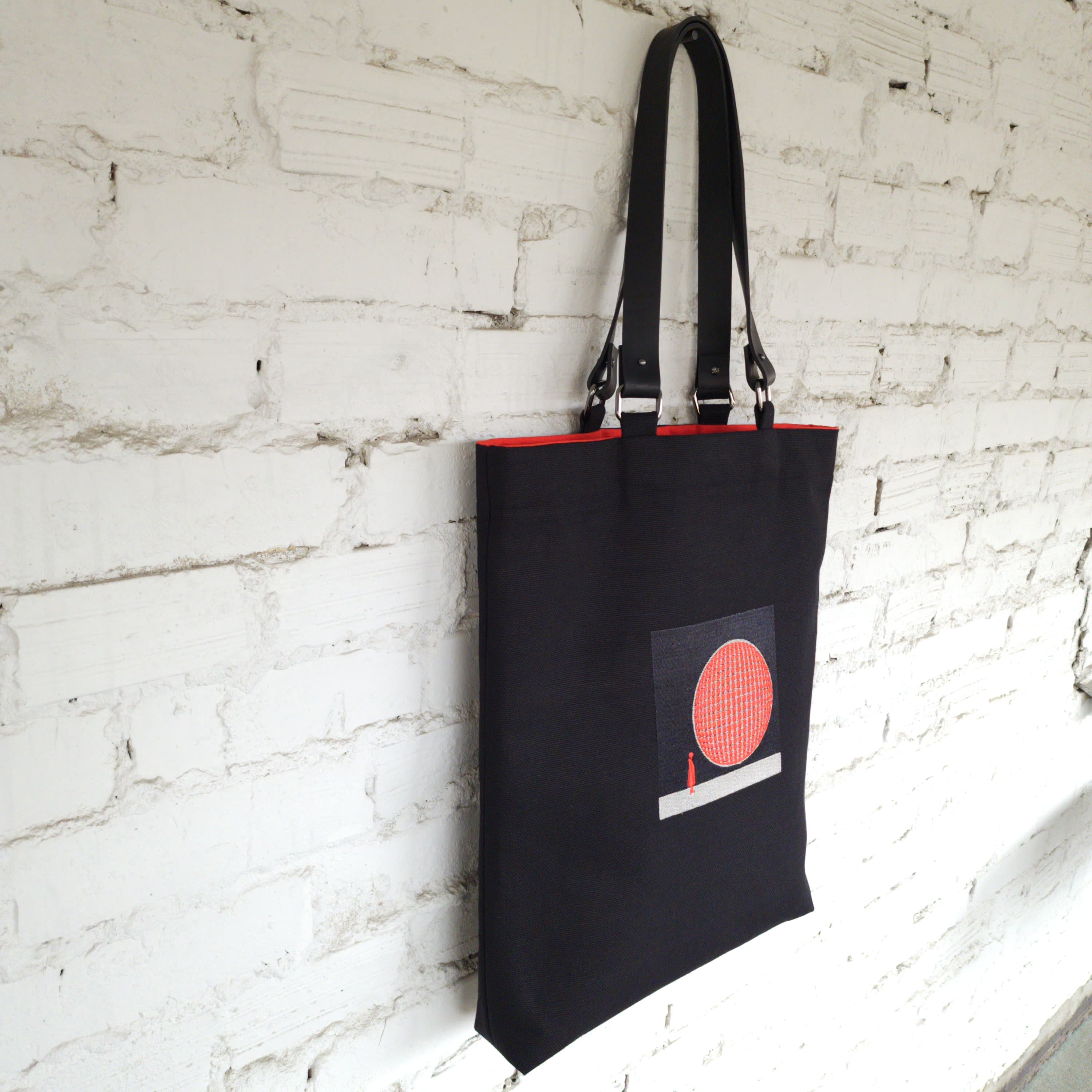 Black linen tote bag with 3D globe embroidery and leather handles, hanging against a white brick wall