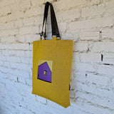 Mustard yellow tote bag with minimalist embroidered purple house design, hanging on white brick wall, shown from side view
