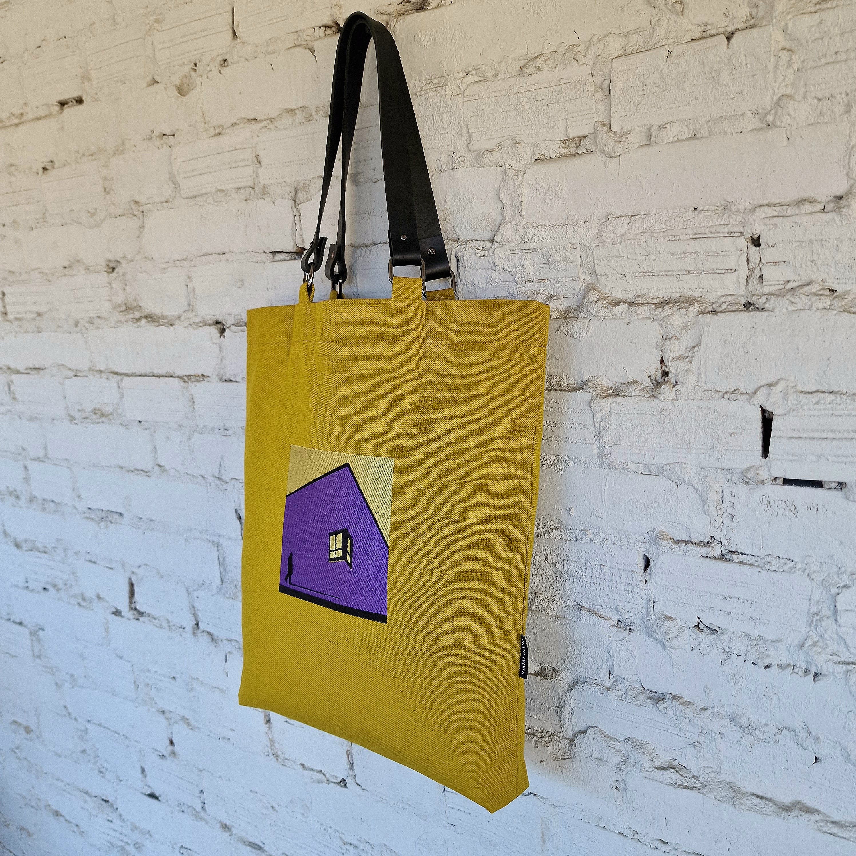 Mustard yellow tote bag with minimalist embroidered purple house design, hanging on white brick wall, shown from side view