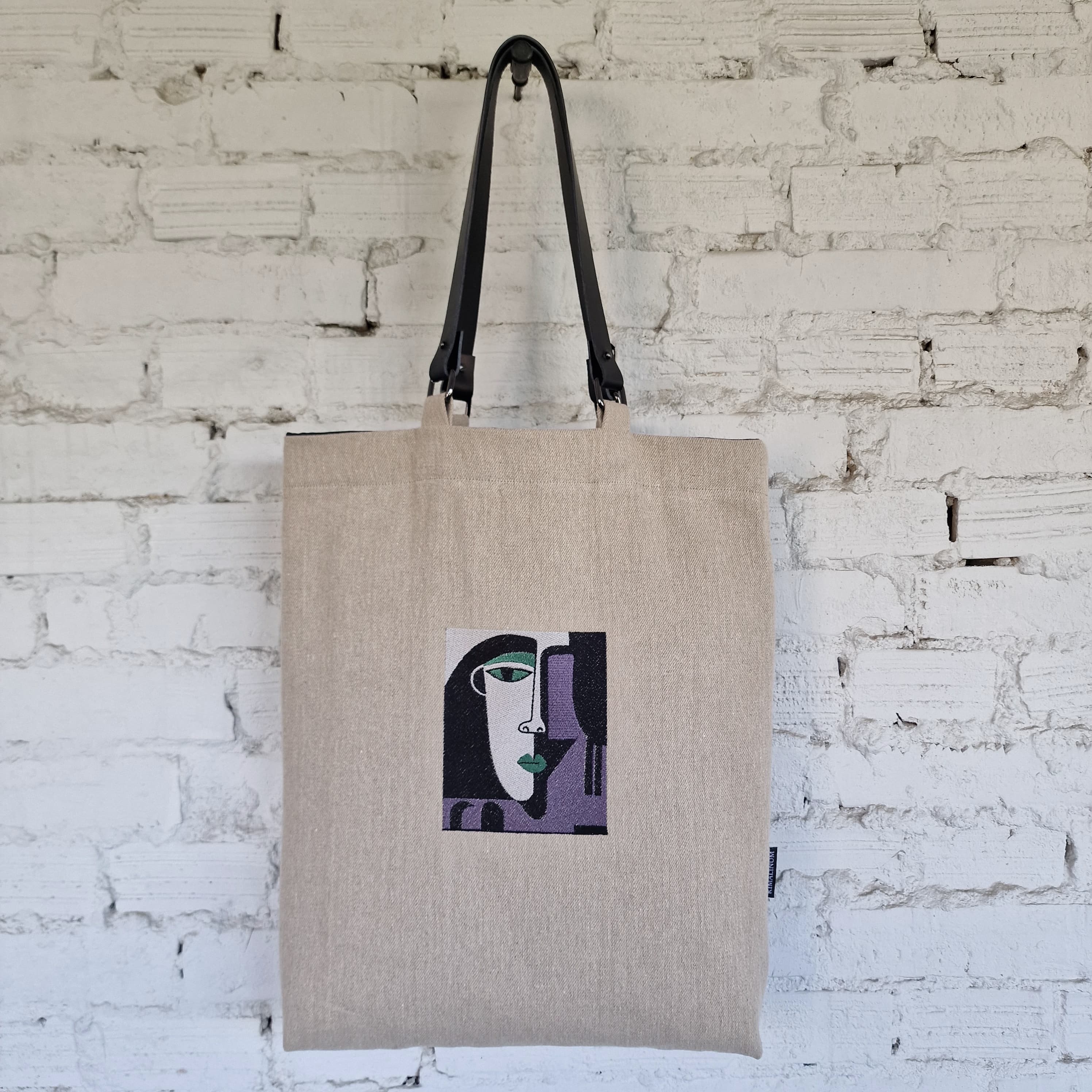 Full front view of large natural linen tote bag with black detachable shoulder straps and unique modern embroidery.