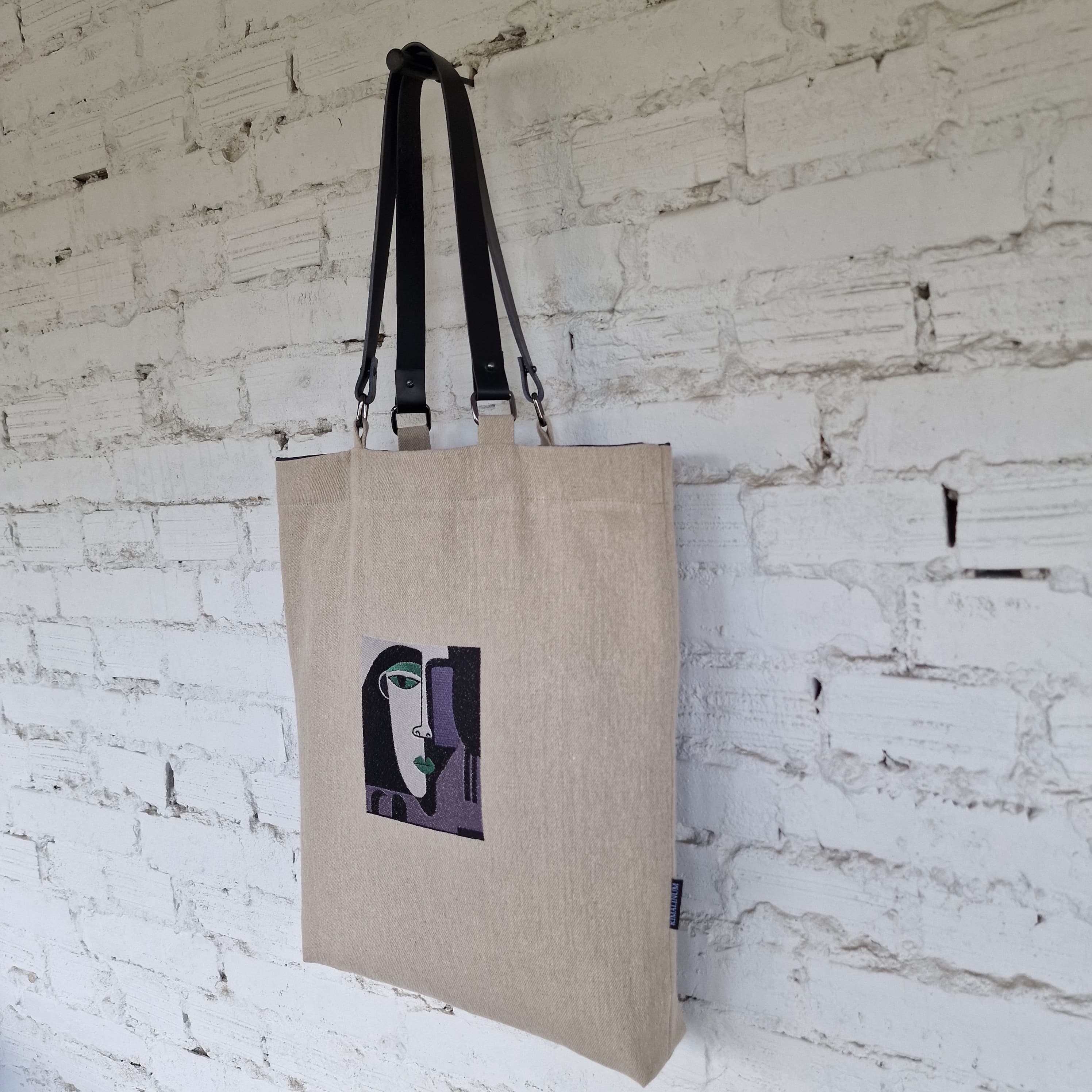 Natural linen tote bag hanging on a white textured wall, showcasing cubist-style embroidered face in green, grey and black tones.