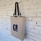 Natural linen tote bag with black leather handles, featuring embroidered abstract face design, photographed from side angle on neutral background.