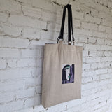 Natural linen tote bag hanging on a white textured wall, showcasing cubist-style embroidered face in green, lilac, and black tones