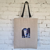 Natural linen tote bag with black leather handles and embroidered abstract face, hanging on white textured brick wall in minimalist setting.