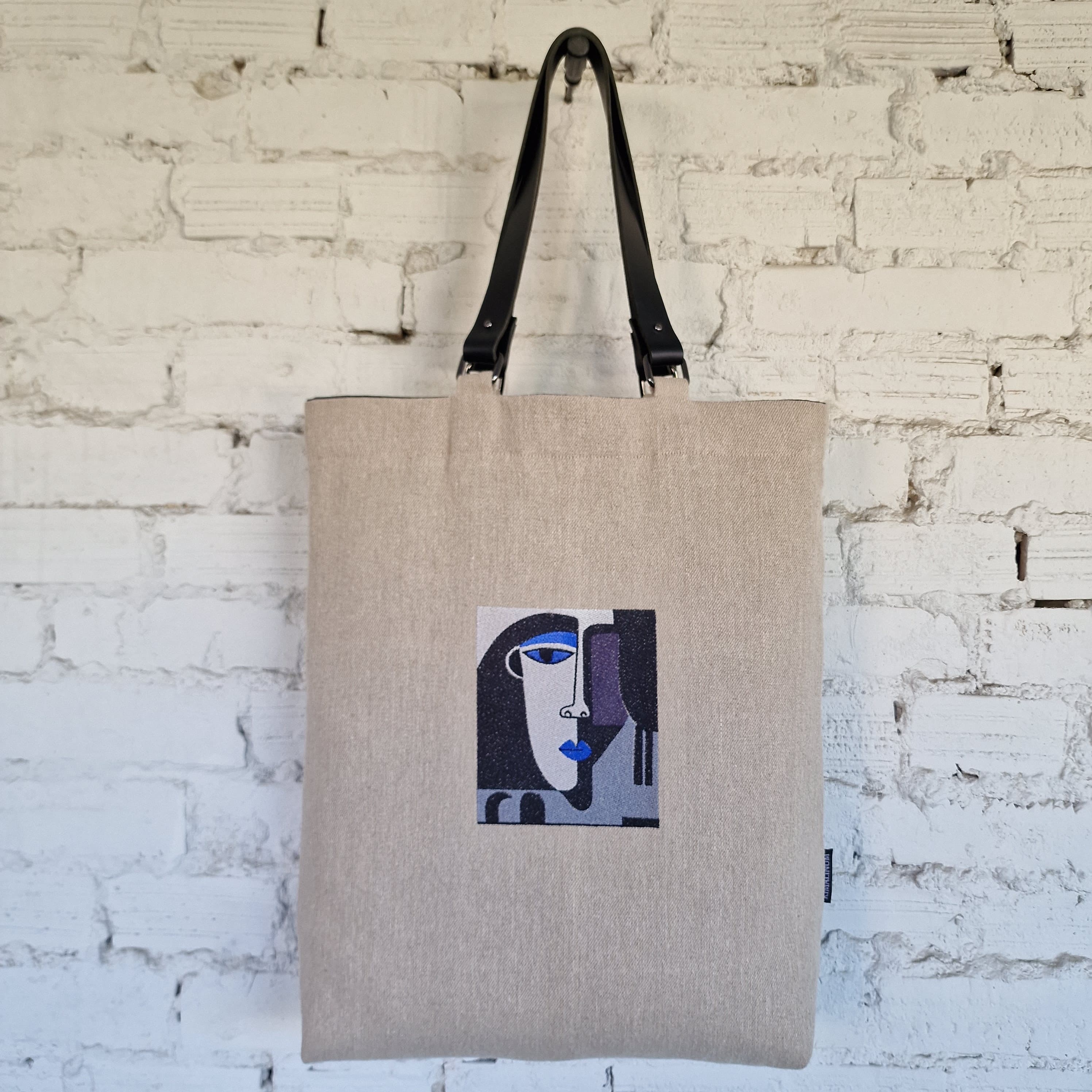 Natural linen tote bag with black leather handles and embroidered abstract face, hanging on white textured brick wall in minimalist setting.
