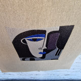 Detailed view of abstract embroidered face design on natural linen tote bag, featuring bold black, grey, and blue stitching on textured fabric.