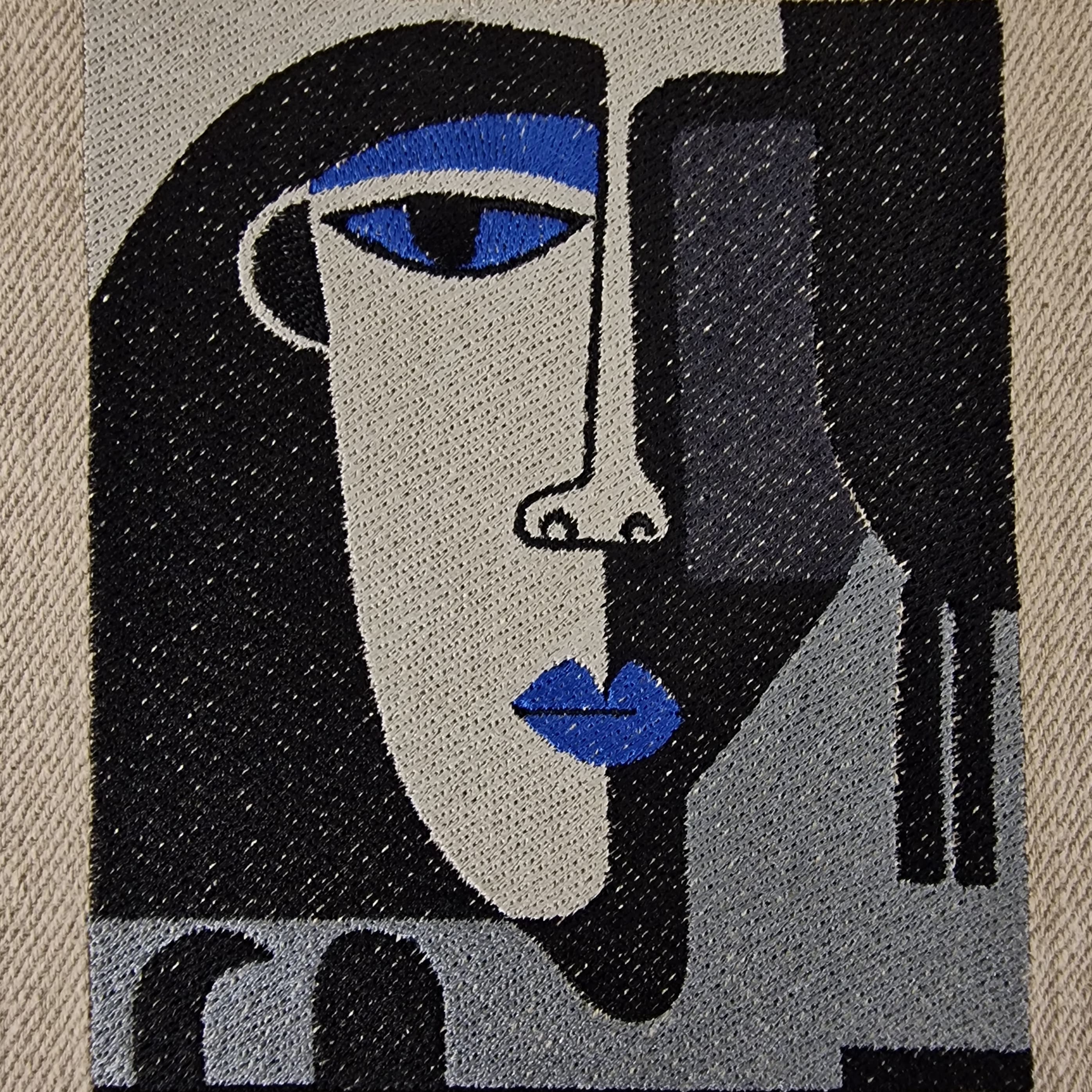 Extreme close-up of embroidered abstract face on natural linen tote bag, showing fine stitching details in black, grey, and bold blue thread.