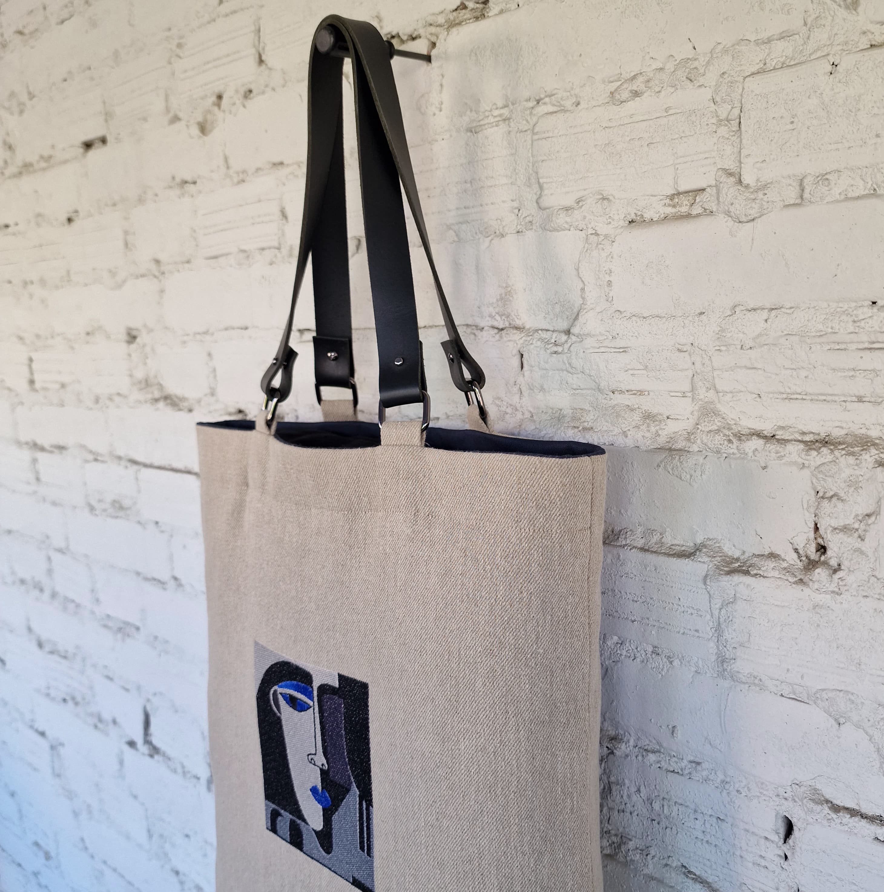 Detailed view of abstract embroidered face design on natural linen tote bag, featuring bold black, grey, and blue stitching on textured fabric.