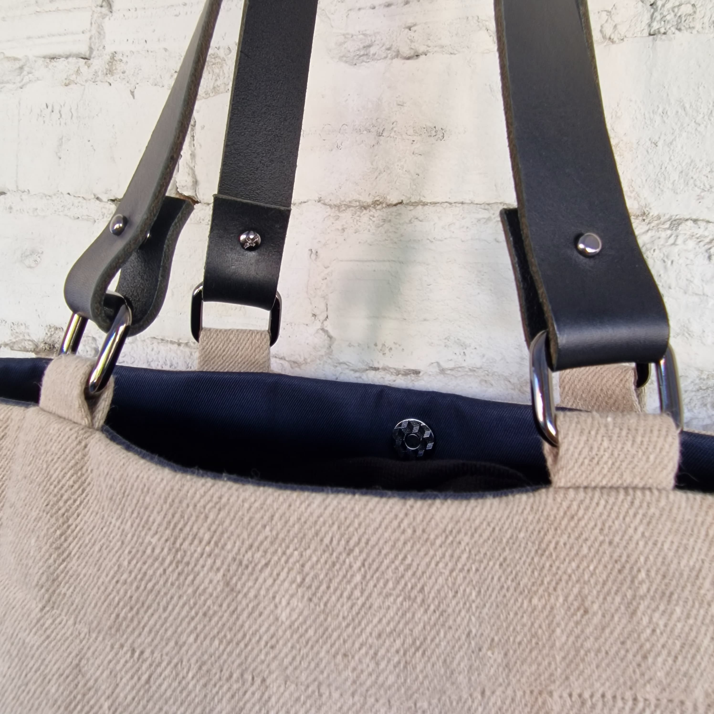 Close-up of black leather tote bag handles attached with metal hardware to natural linen fabric, showing navy cotton lining and magnetic snap closure.
