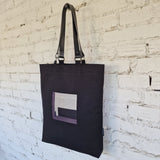 Angled side view of black tote bag with embroidered square detail and leather straps