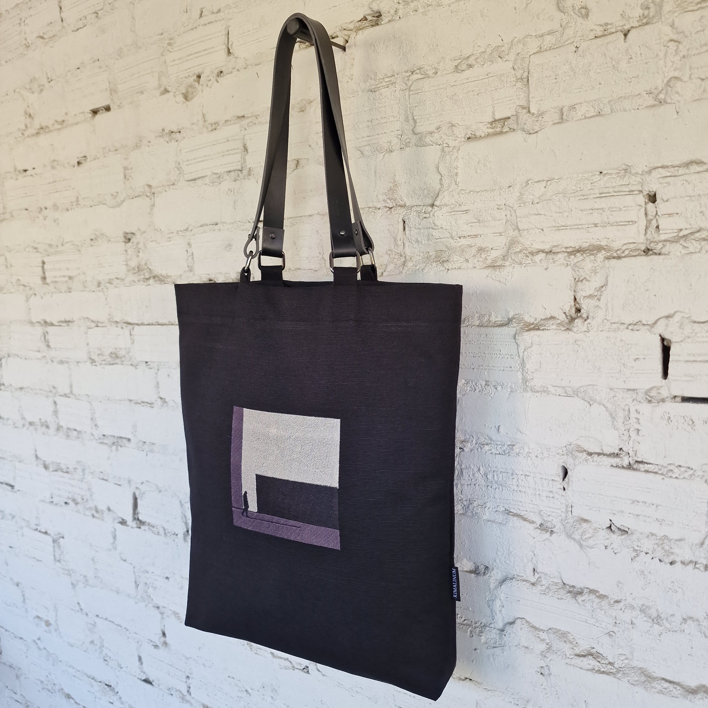 Angled side view of black tote bag with embroidered square detail and leather straps