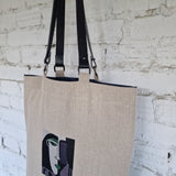 Minimalist natural linen tote bag hanging against a white brick wall, featuring bold abstract face embroidery in modern cubist style.