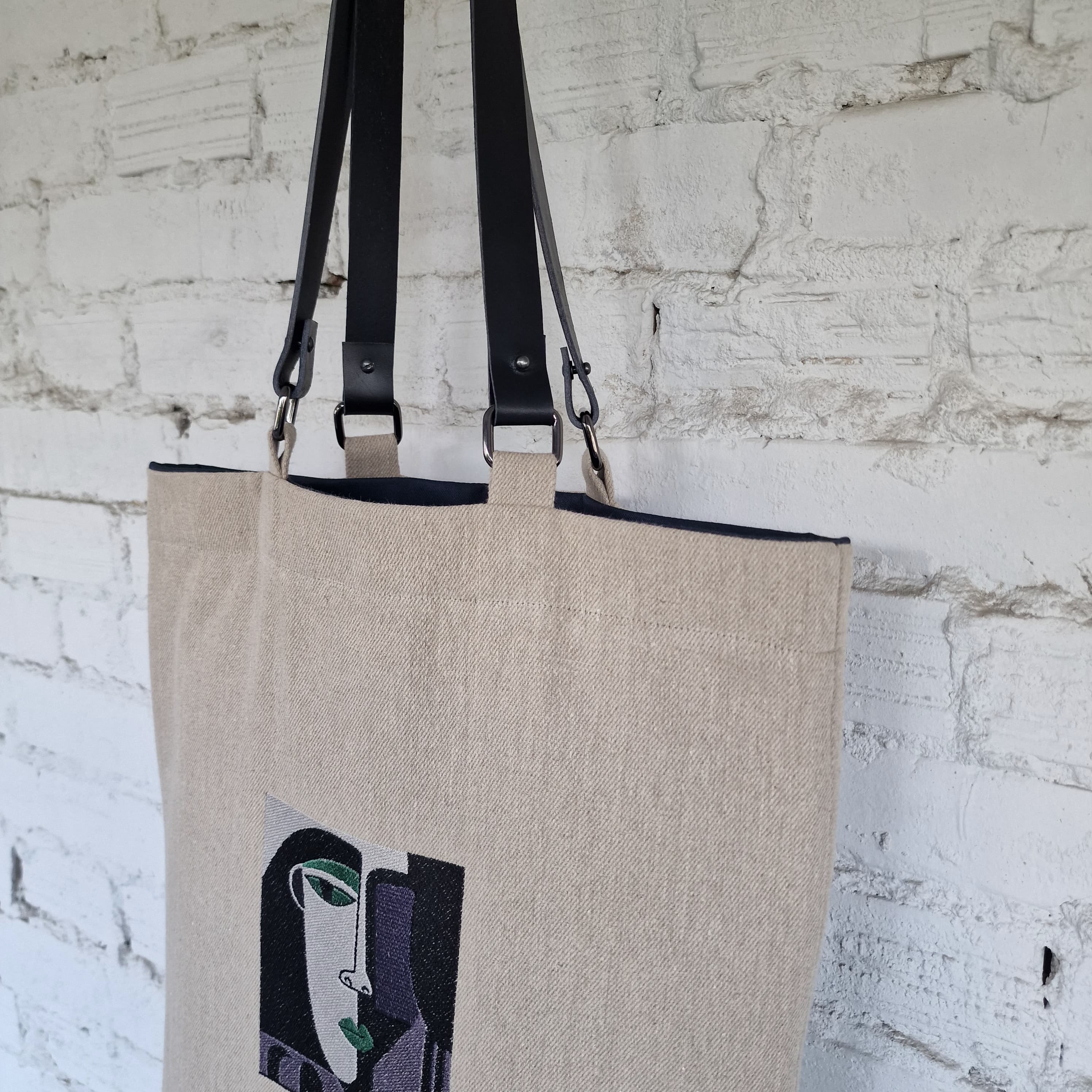 Minimalist natural linen tote bag hanging against a white brick wall, featuring bold abstract face embroidery in modern cubist style.
