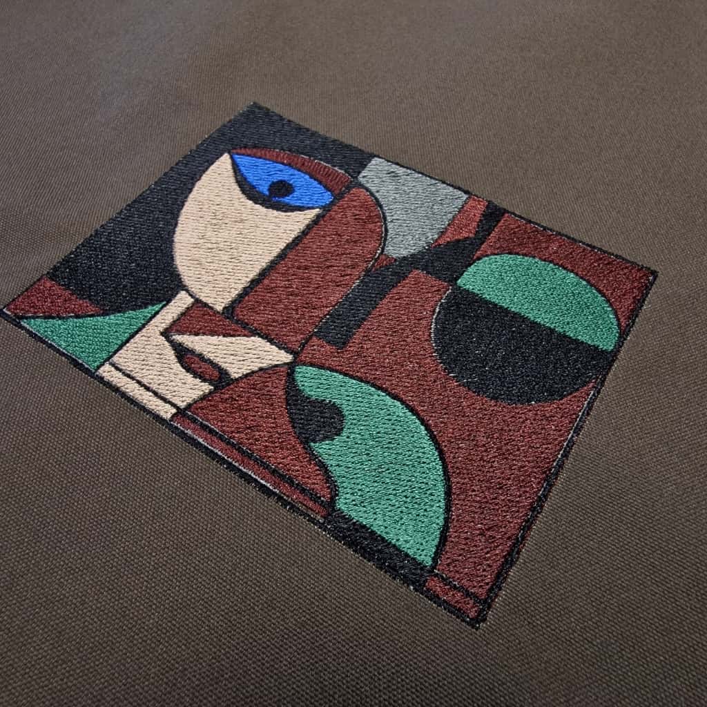 Macro shot of abstract face embroidery in red, green, beige, and blue thread on dark brown cotton fabric – RIMALINUM tote bag detail
