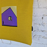 Mustard yellow tote bag with black leather handles and embroidered purple house design, hanging on white textured wall