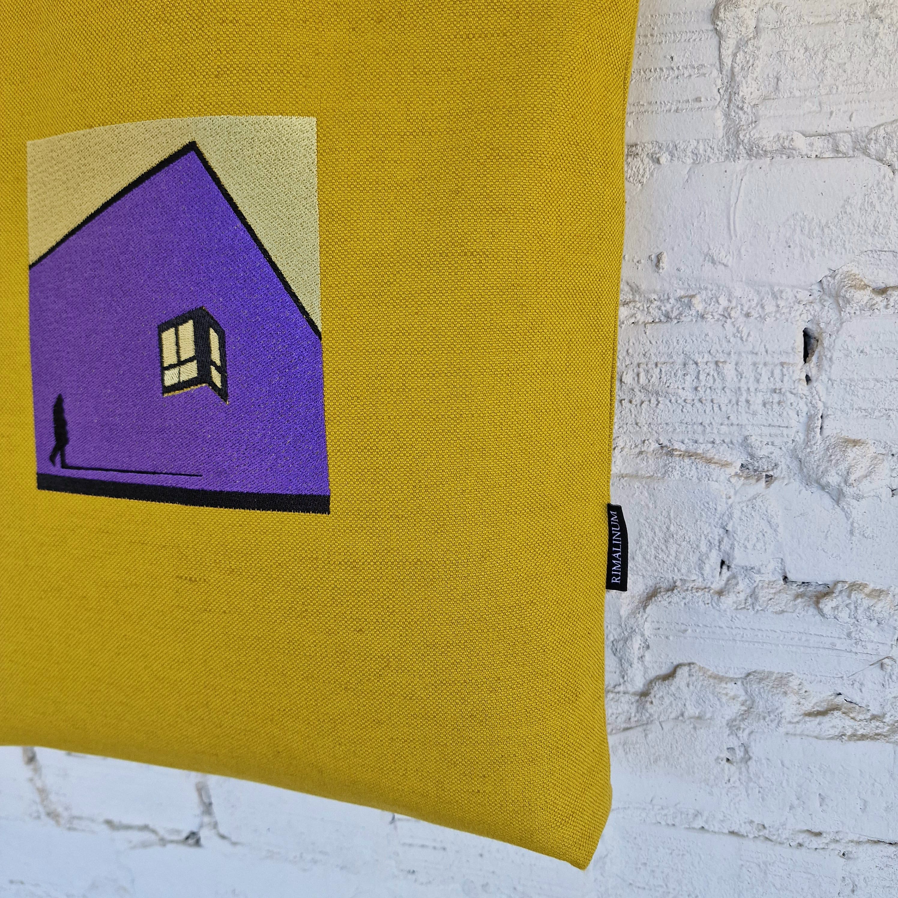 Mustard yellow tote bag with black leather handles and embroidered purple house design, hanging on white textured wall