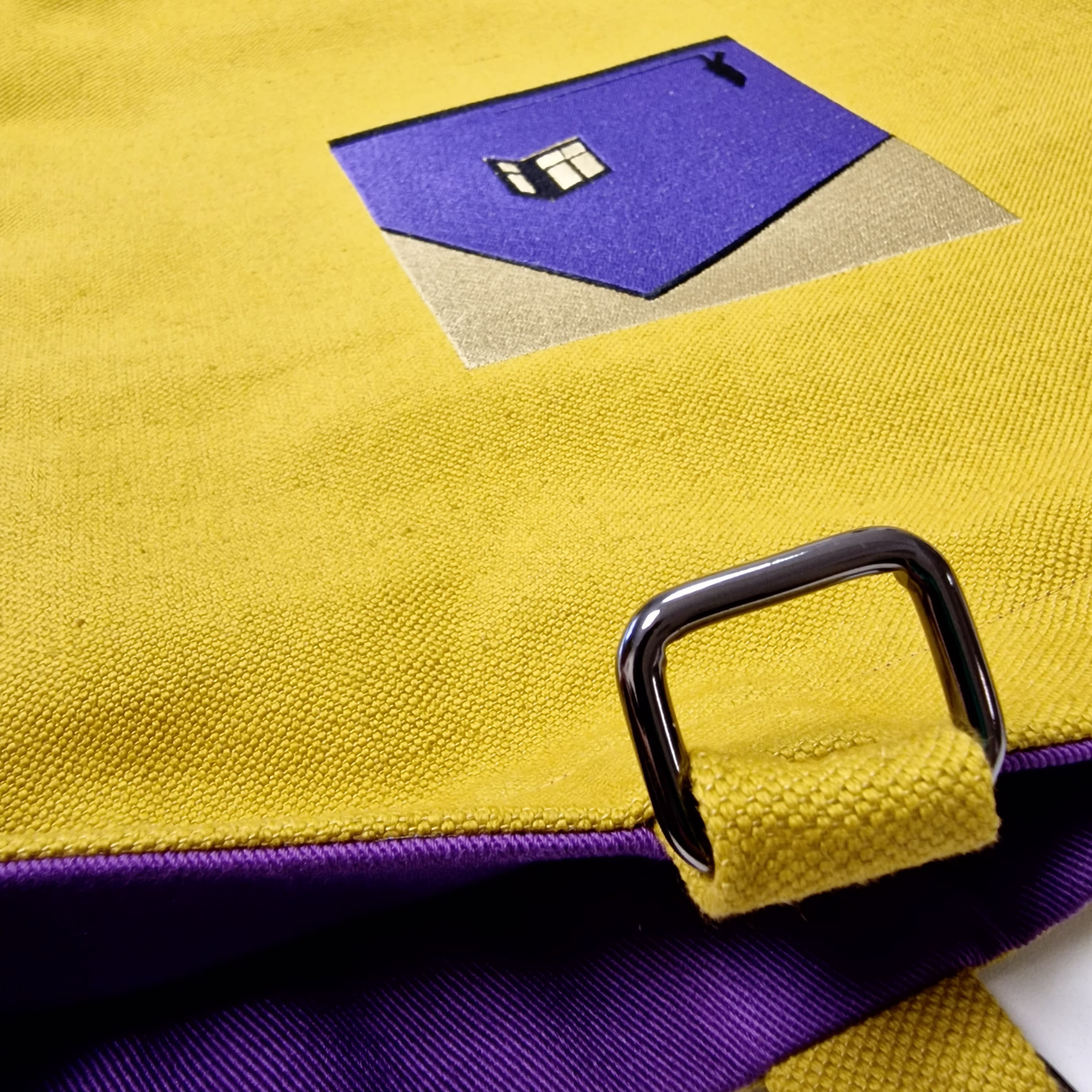 Close-up of yellow canvas fabric with purple embroidered house design, visible purple lining and silver metal hardware detail