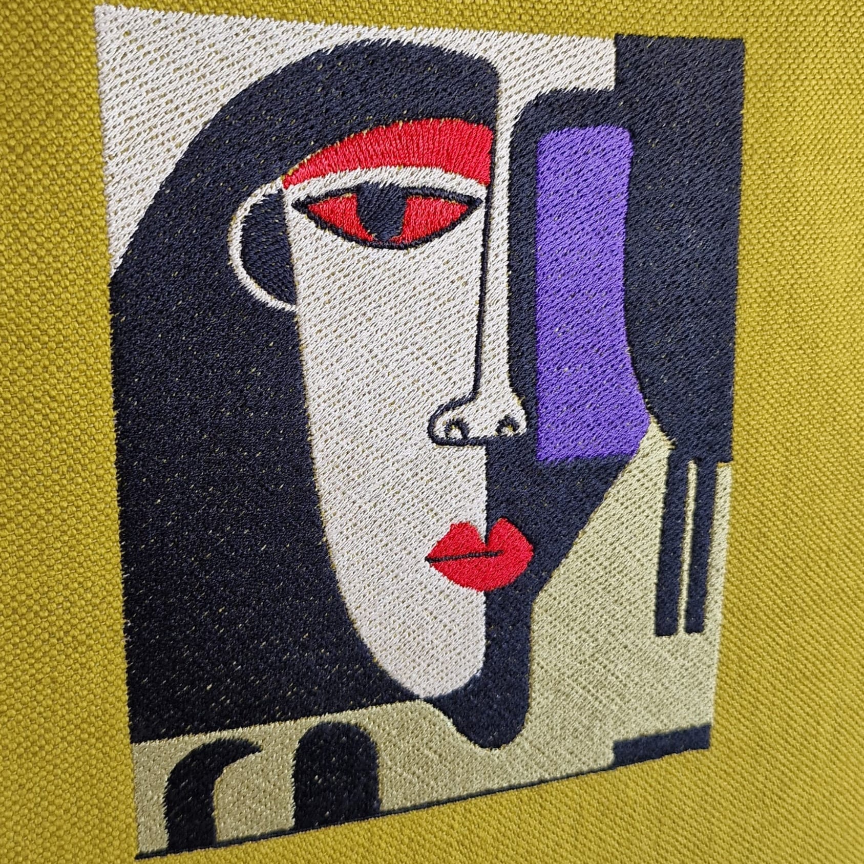 Close-up of abstract embroidered face in red, purple, cream, and black thread on mustard yellow linen fabric, showing fine stitch details