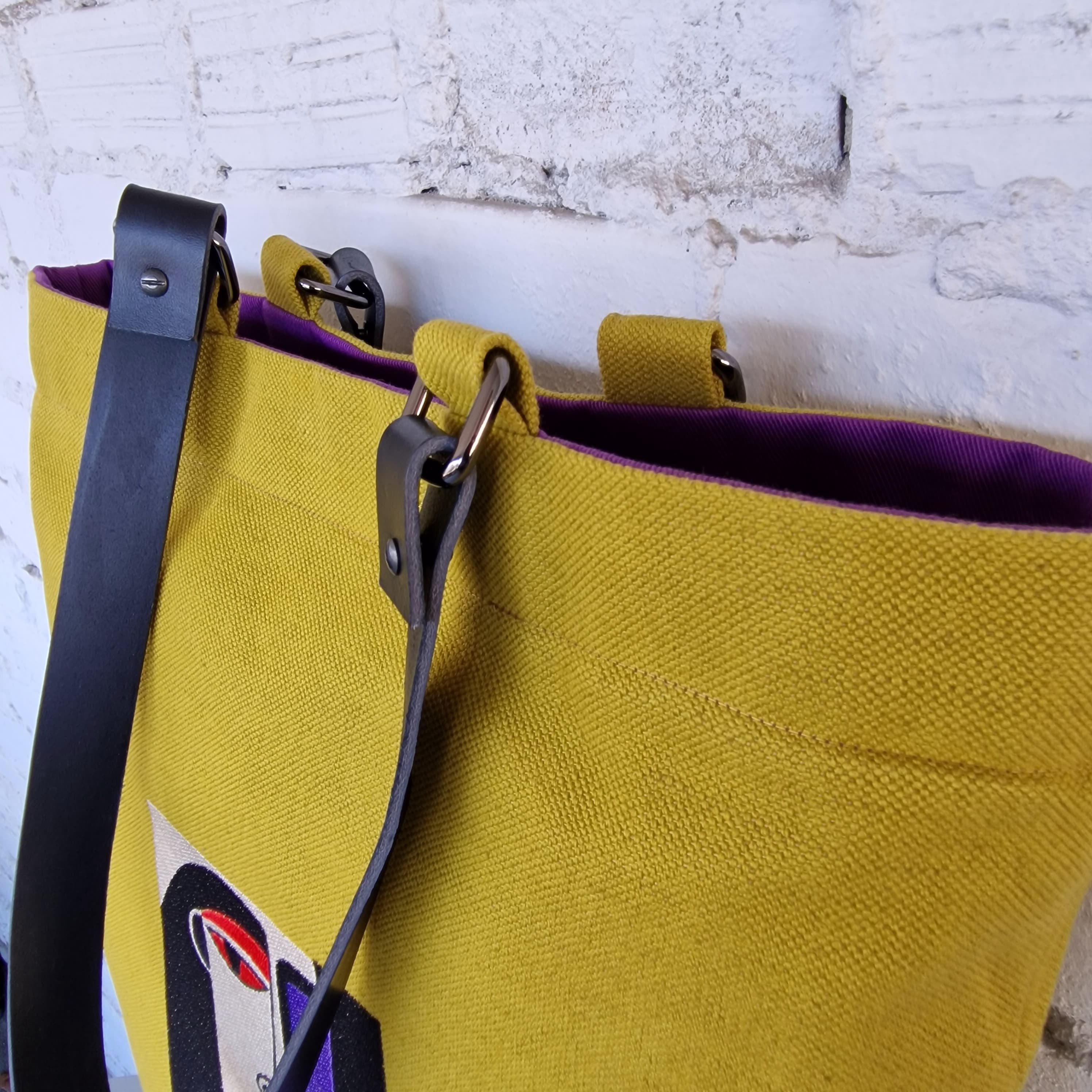 Close-up of mustard yellow linen tote bag with black leather straps and purple cotton lining, showing metal ring attachments and stitching details