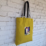 Yellow tote bag with a graphic design of a face on a white brick wall.