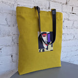 Yellow tote bag with a face design on a white brick wall background
