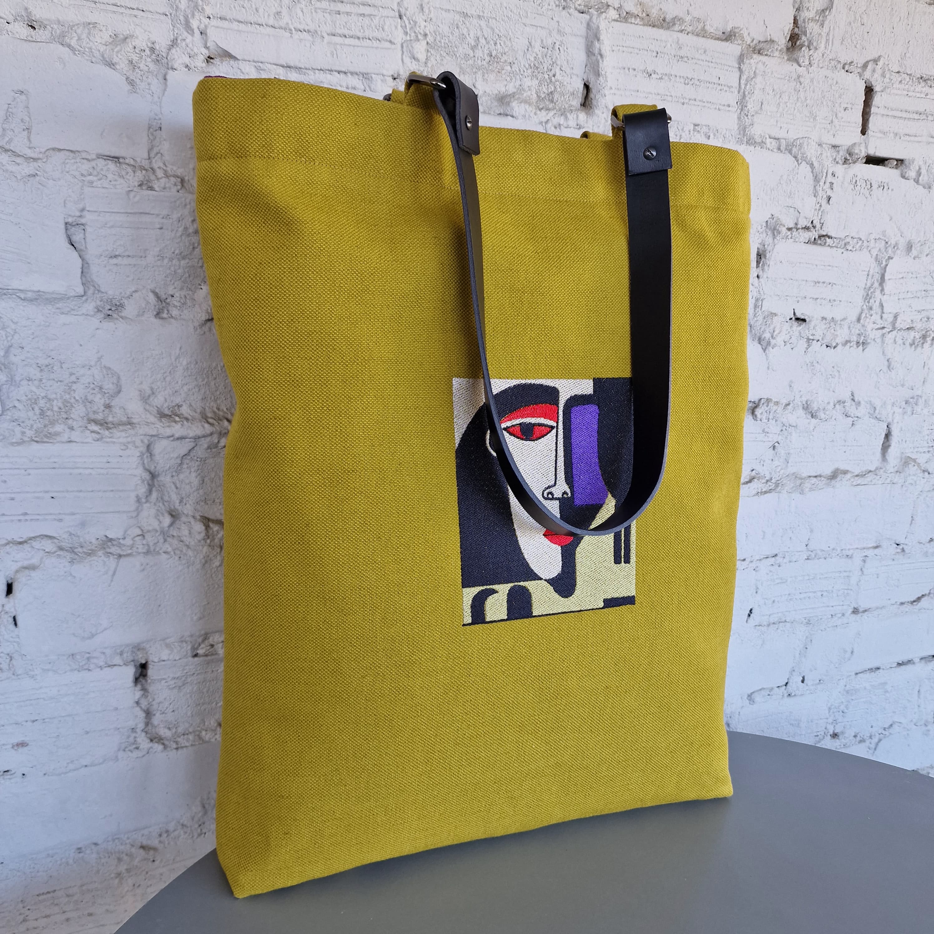 Yellow tote bag with a face design on a white brick wall background