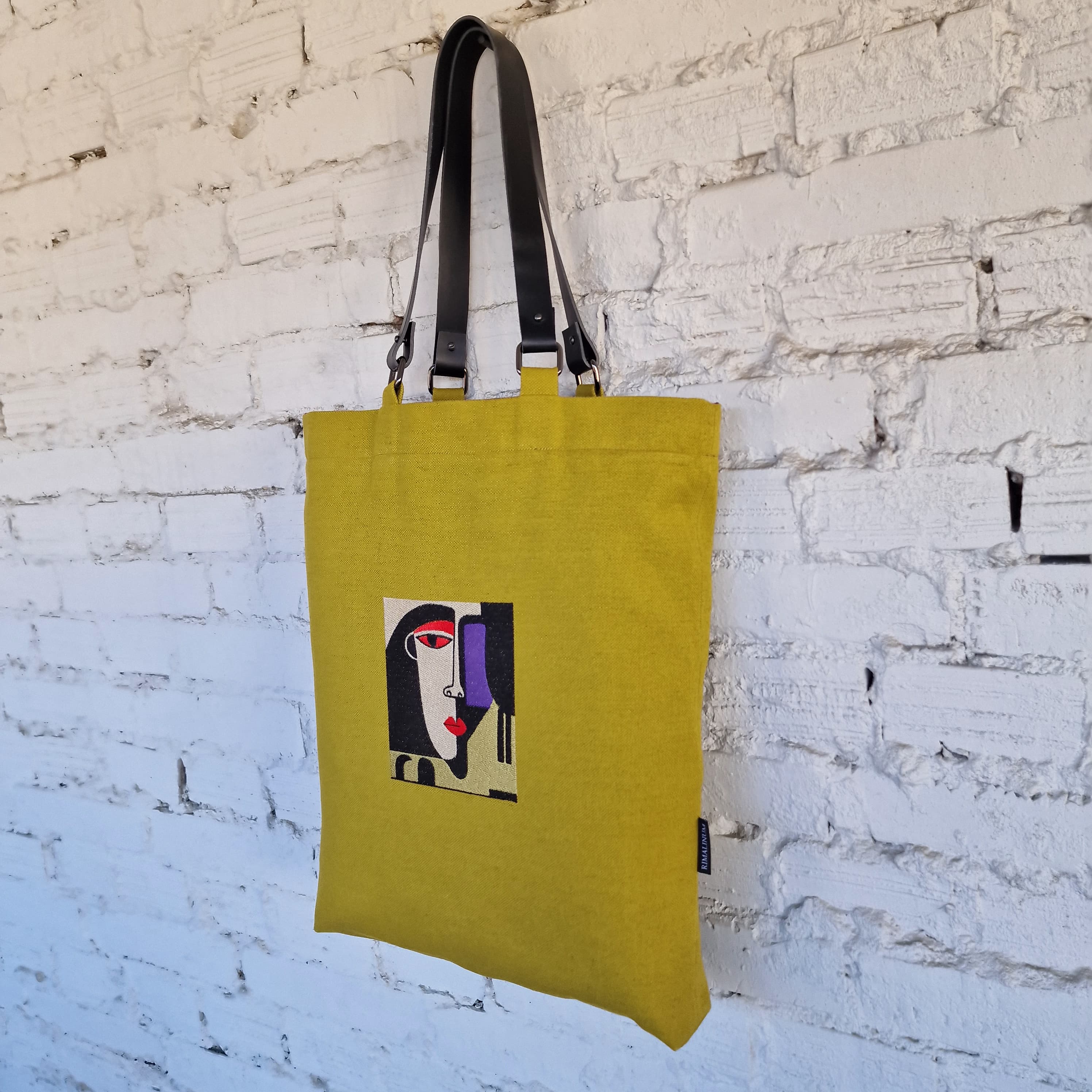 Yellow tote bag with a graphic design against a white brick wall
