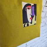 Detailed view of abstract embroidered face design on mustard yellow linen tote, with red, black, cream, and purple thread on textured fabric