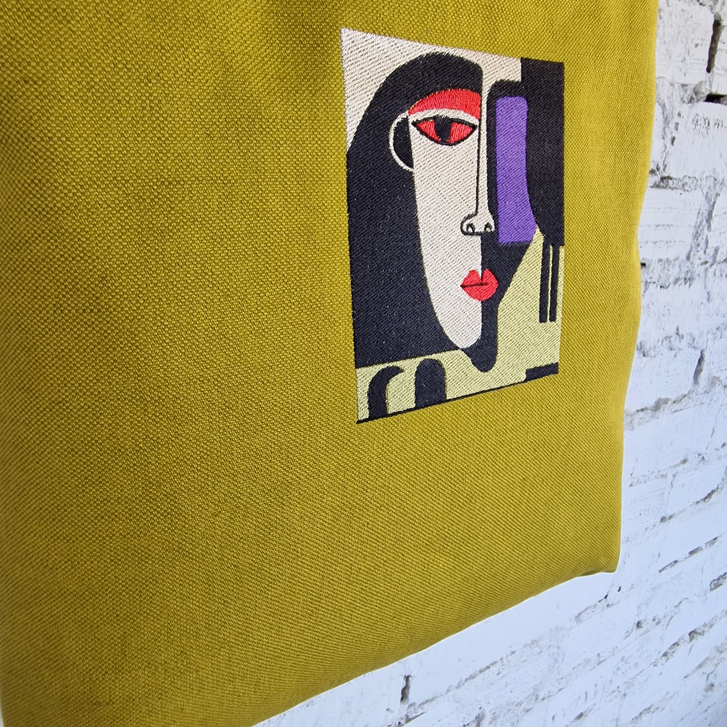 Detailed view of abstract embroidered face design on mustard yellow linen tote, with red, black, cream, and purple thread on textured fabric