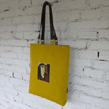 Yellow tote bag with a graphic design of a face on a white brick wall.
