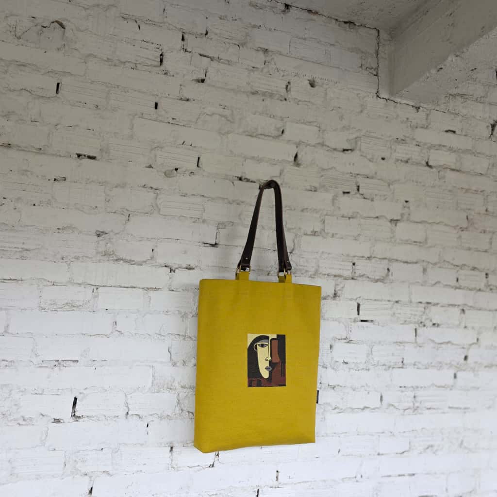 Yellow tote bag with a graphic design hanging on a white brick wall.