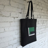 Black linen tote bag with green embroidered minimalist design and removable leather handles hanging on a white brick wall