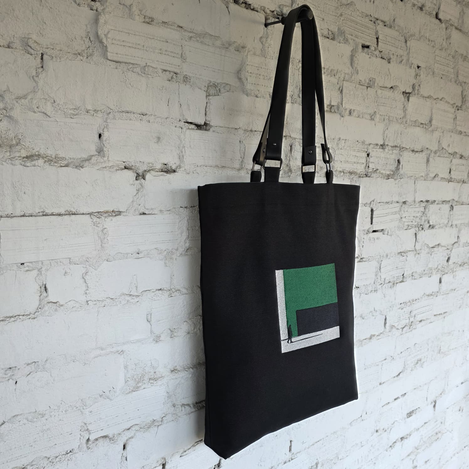 Black linen tote bag with green embroidered minimalist design and removable leather handles hanging on a white brick wall