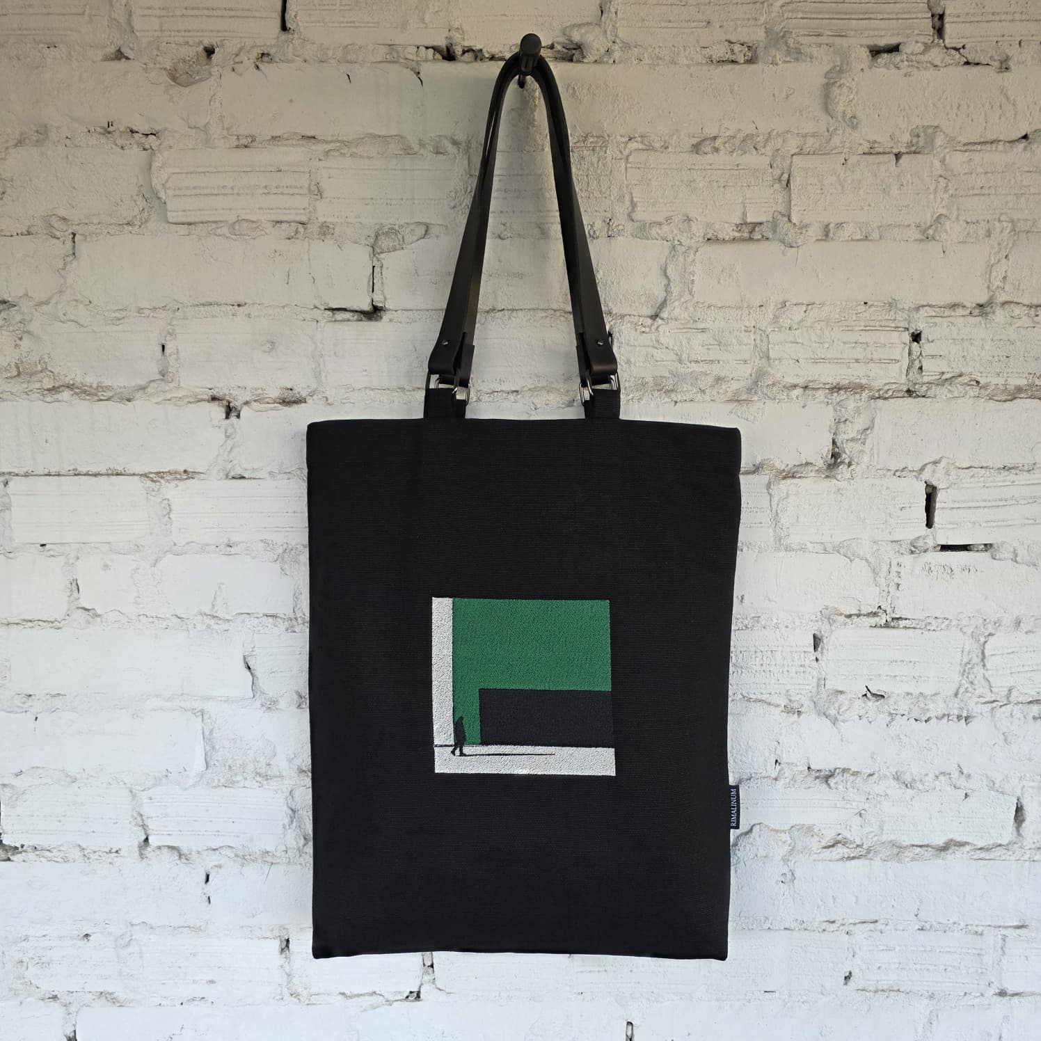 Front view of black linen tote bag featuring green embroidered art and removable leather handles, displayed on a white brick wall