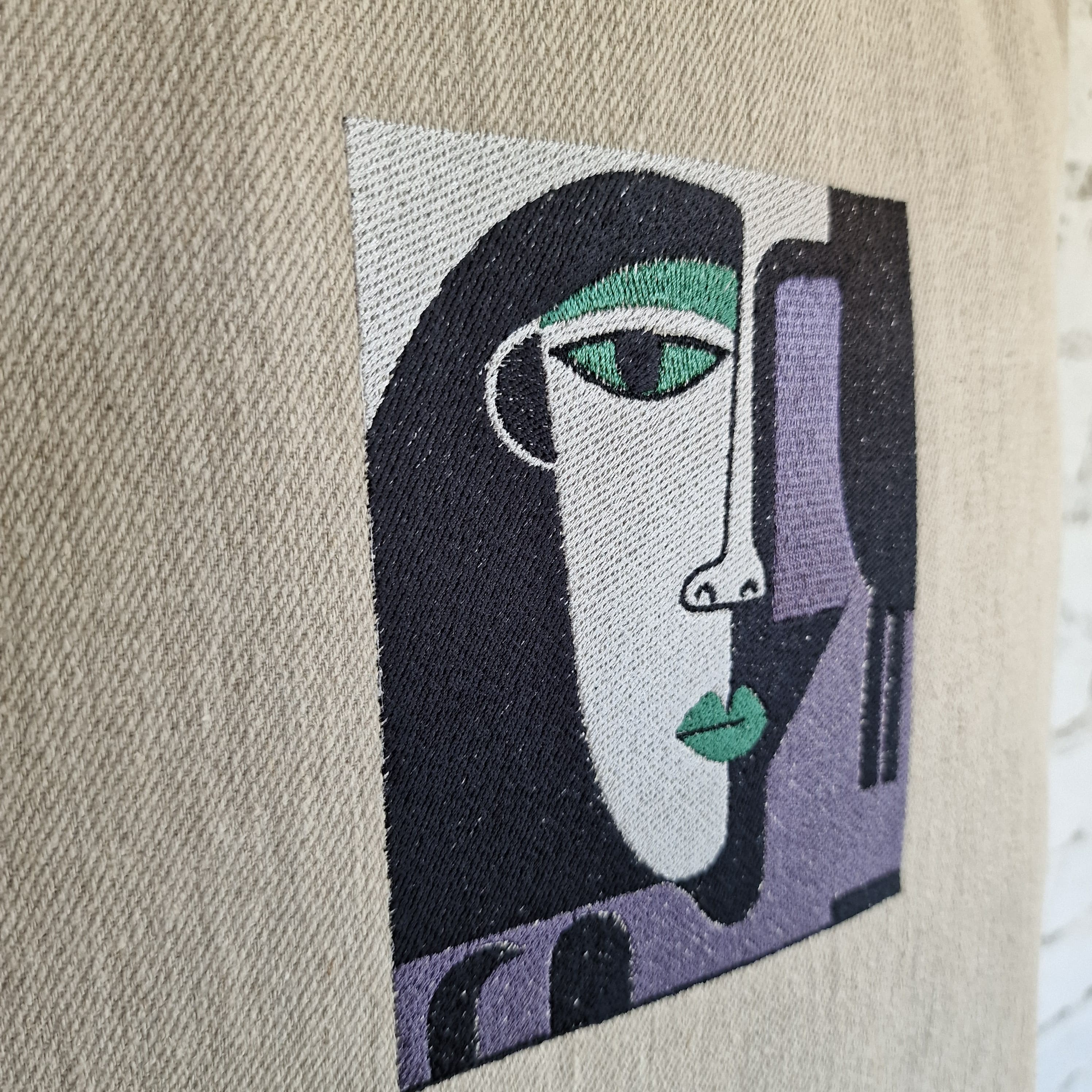 Macro detail of geometric cubist-style embroidered face – green eye, structured forms, and stitched texture on beige linen fabric