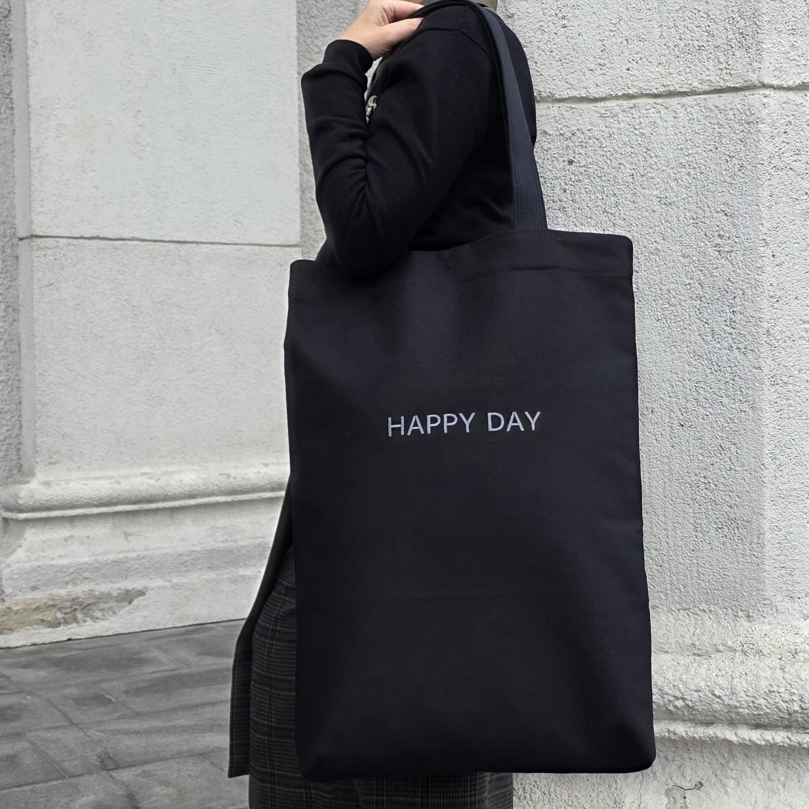 Person holding a black tote bag with 'HAPPY DAY' text against a stone wall.