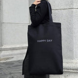 Person holding a black tote bag with 'HAPPY DAY' text against a stone wall.