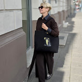 A person walking on a city sidewalk carrying a large black tote bag with a graphic print. The person is wearing a dark brown coat, dark pants, and sunglasses.