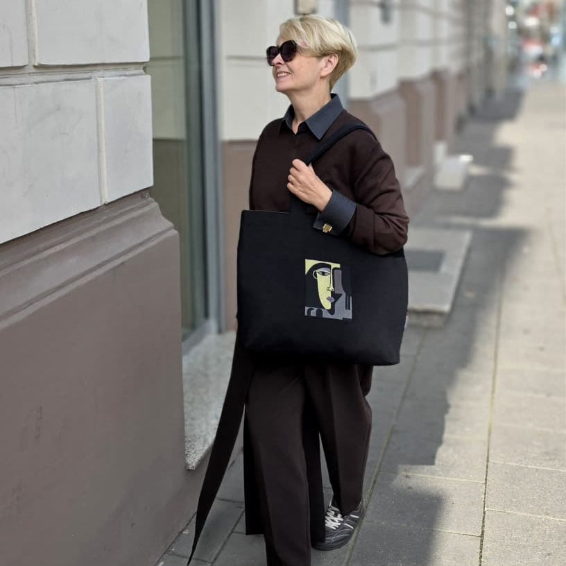 A person walking on a city sidewalk carrying a large black tote bag with a graphic print. The person is wearing a dark brown coat, dark pants, and sunglasses.