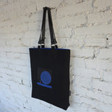 Black tote bag with a blue graphic design hanging against a white brick wall.