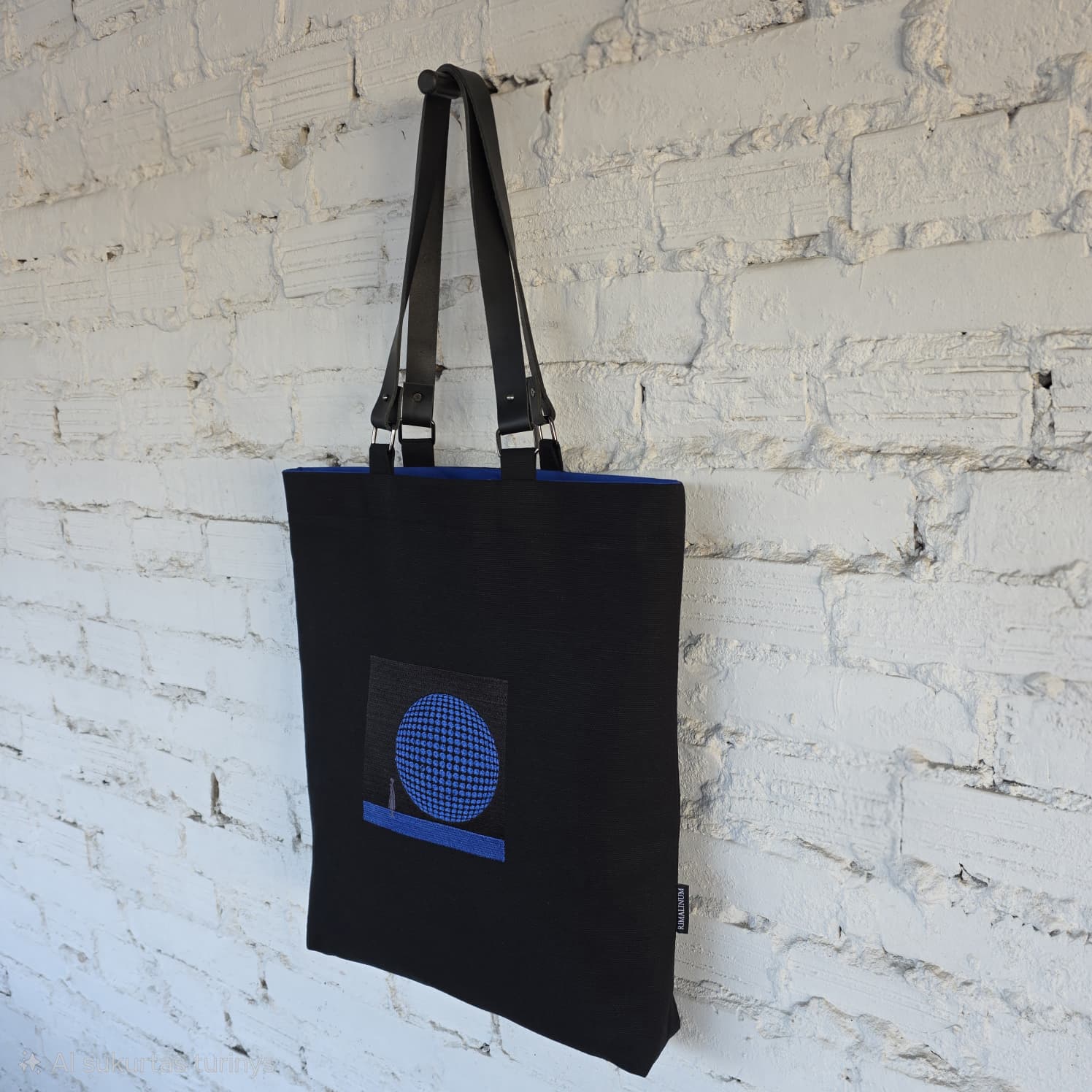 Black tote bag with a blue graphic design hanging against a white brick wall.