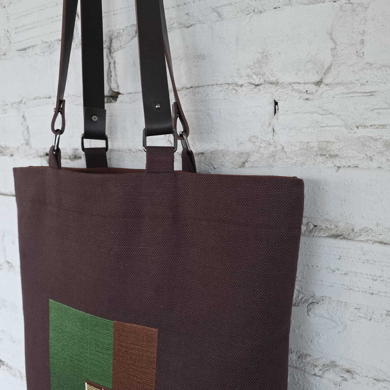 Brown tote bag with green and brown square design against a white brick wall.